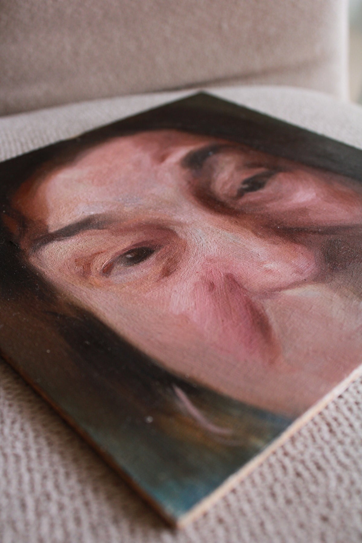 A close-up photograph of a painted portrait of a person's face, shown resting on a textured beige fabric surface.